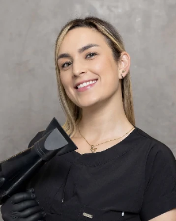 Catherine Cadavid con cabello rubio liso y un secador en la mano posando con una gran sonrisa mientras usa una chaqueta negra